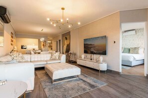 A modern living room with a sectional sofa, a coffee table, a TV, and a painting on the wall. - Houghton Views - Camps Bay Apt, Pool, Ocean Views (Cape Town)