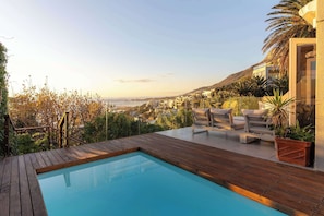 Un área de piscina con vistas a una ciudad costera, terraza de madera y mobiliario exterior.