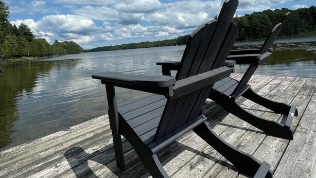 A wooden dock with two Adirondack chairs facing a calm lake, surrounded by trees under a partly cloudy sky.