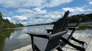 Une jetée en bois avec deux chaises Adirondack, donnant sur un lac paisible, entourée d’arbres sous un ciel partiellement nuageux.