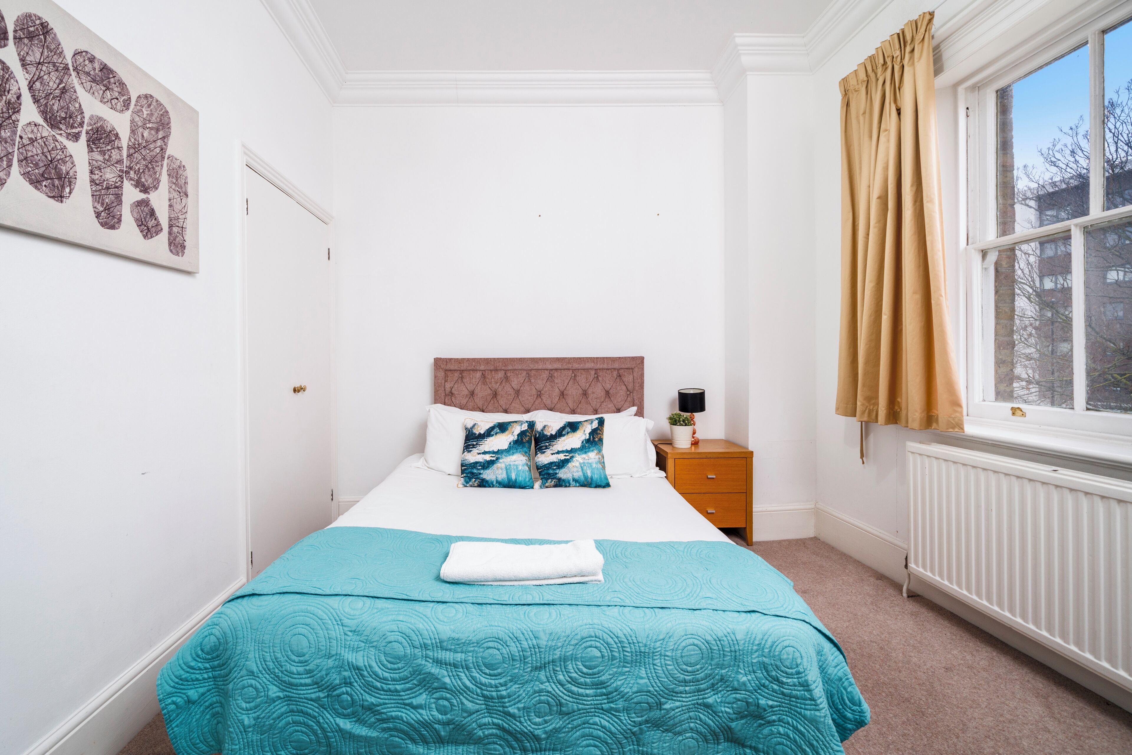 A neatly arranged bedroom with a bed, a bedside table with a lamp, a window with curtains, and a wall with a framed picture.