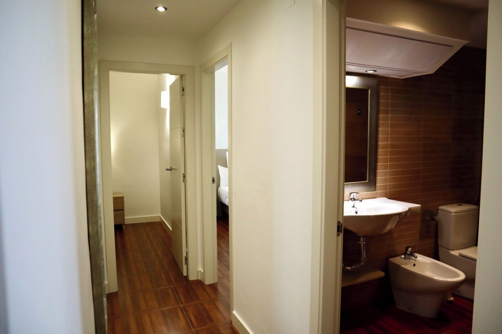 A narrow hallway leading to a bathroom with a sink, bidet, and a mirror.