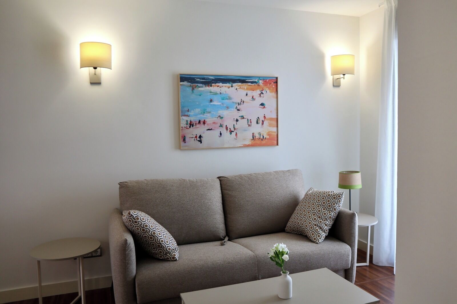 A modern living room with a beige sofa, patterned cushions, a small side table, and a painting of a beach scene.