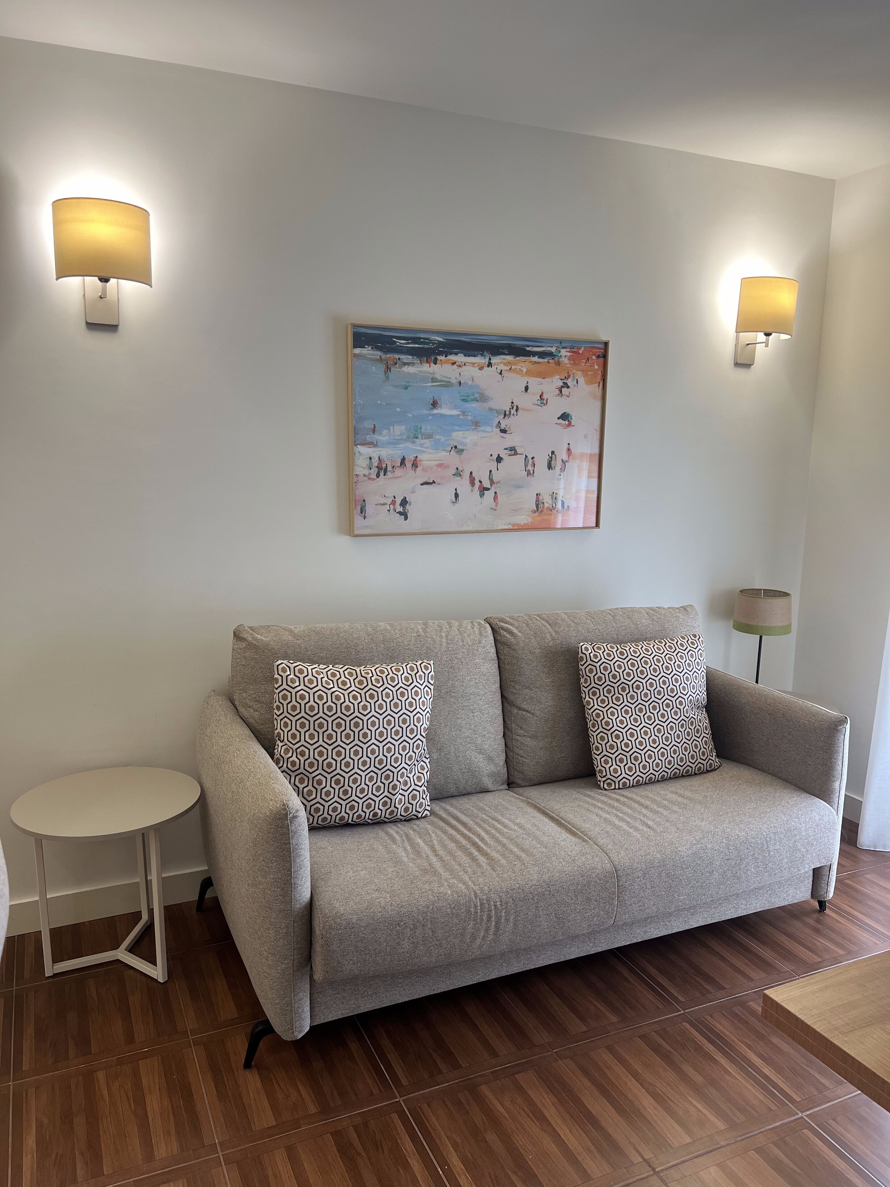 A living room with a grey sofa, patterned cushions, a round side table, and a painting on the wall.