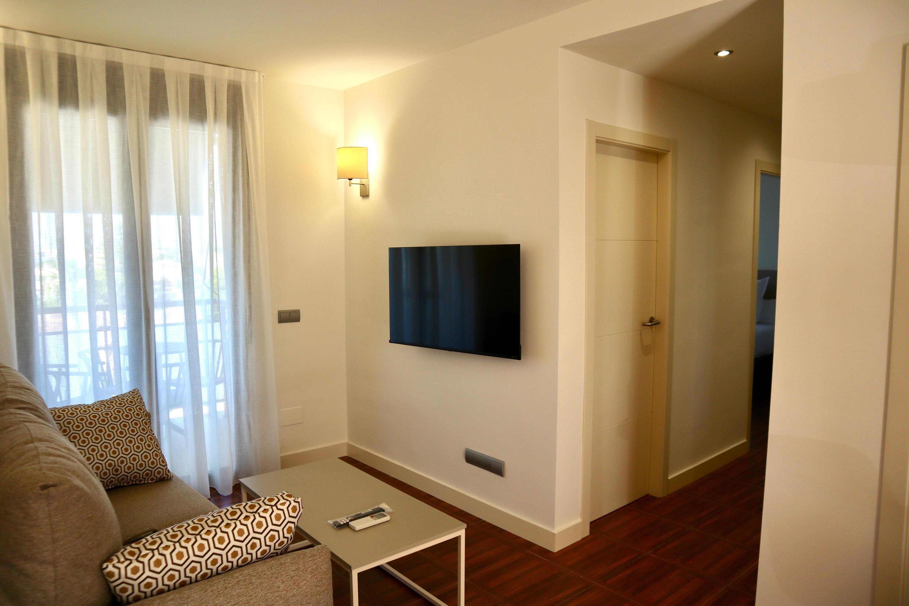 A modern living room with a sofa, patterned pillows, a coffee table, a flat-screen TV mounted on the wall, and a door leading to another room.