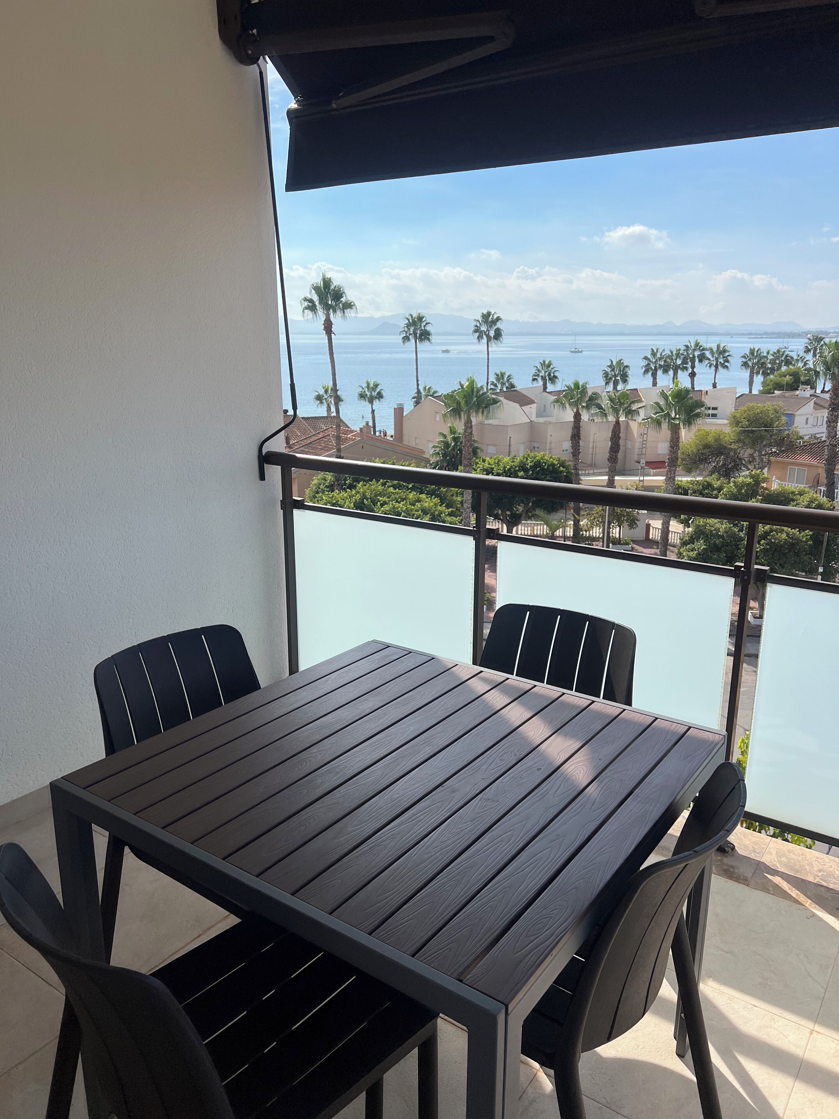 A balcony with a table and chairs, overlooking a coastal area with palm trees and buildings.