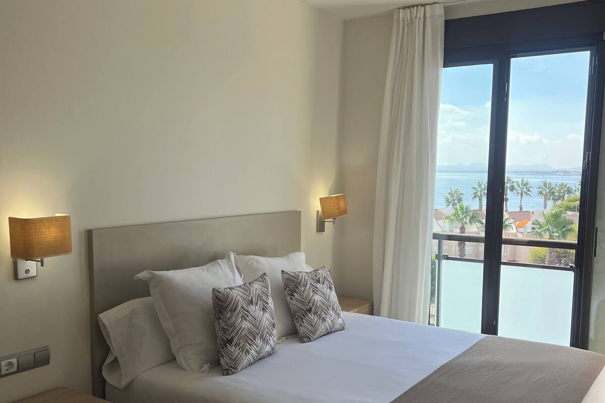 A hotel room with a large bed, two nightstands, a window with a view of the sea, and a ceiling-mounted smoke detector.