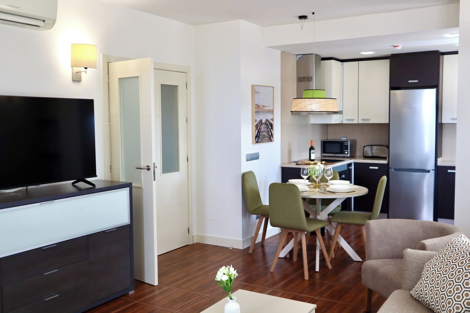 Una sala moderna con televisor de pantalla plana, comedor con mesa lista para dos personas y cocina con electrodomésticos de acero inoxidable.