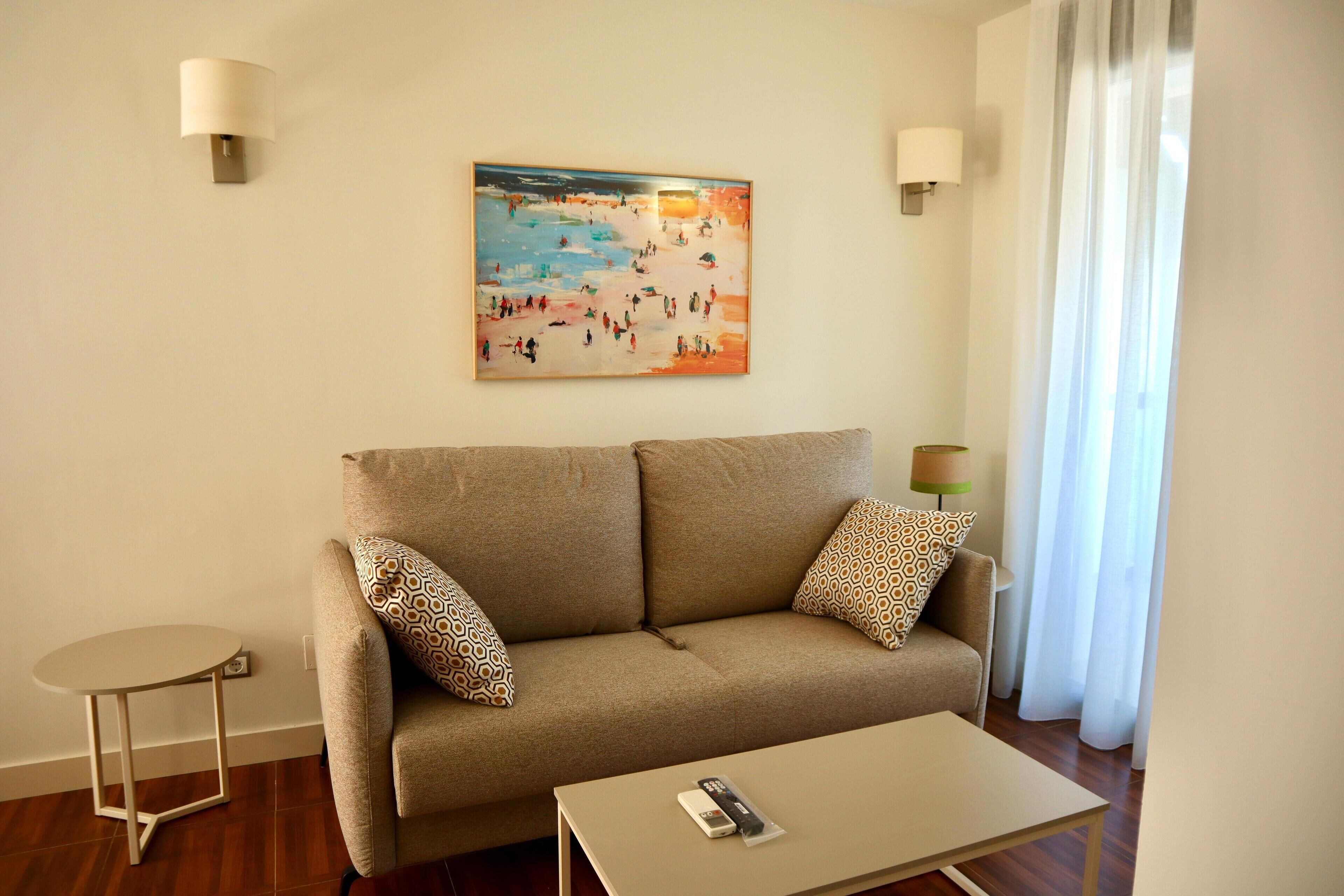 A living room with a beige sofa, patterned pillows, a small round table, and a painting on the wall.