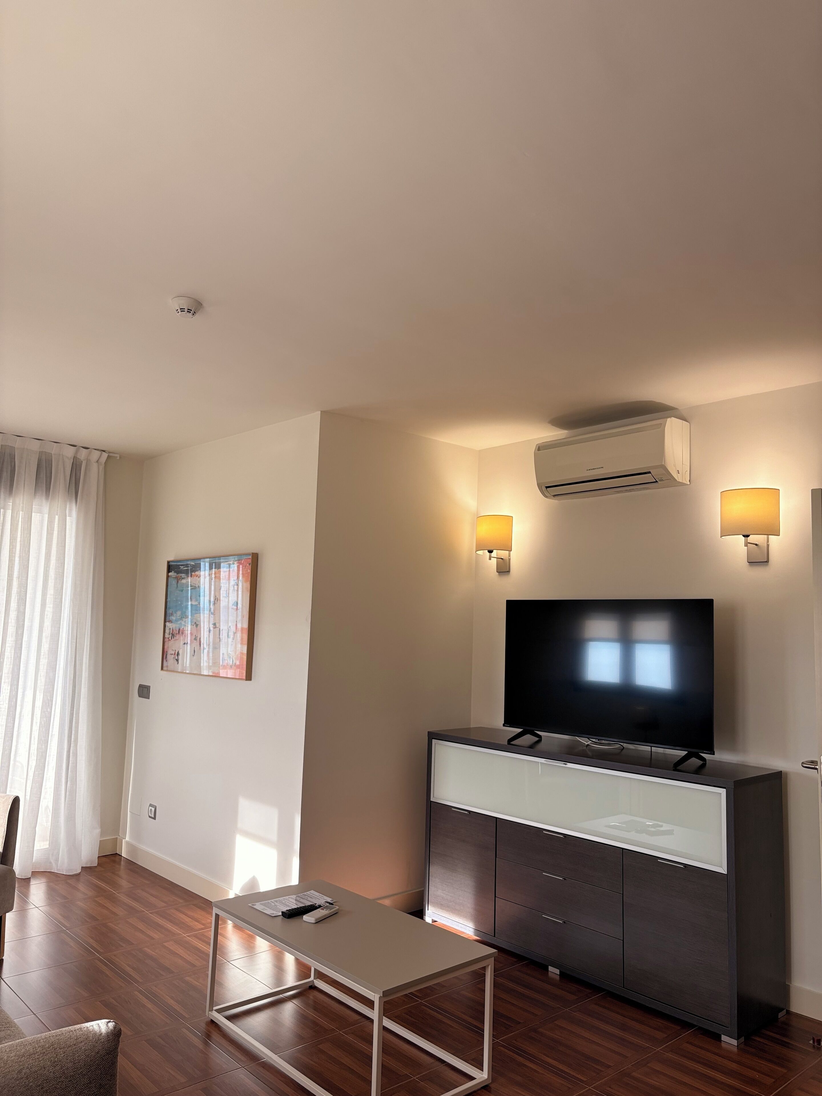 A modern living room with a flat-screen TV, a glass-top coffee table, a dark wooden cabinet, and two wall-mounted lamps.