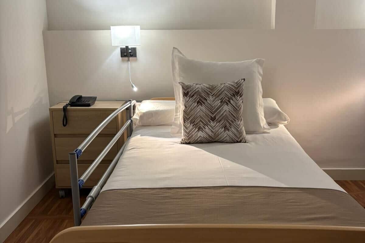 A single bed with a wooden frame, a bedside table with a phone, and a wall-mounted light.