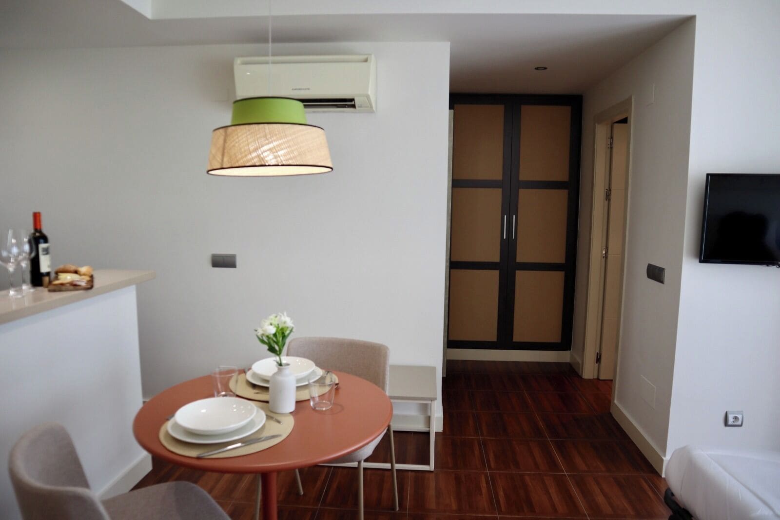 Een knus ingerichte kamer met een eettafel, stoelen, een kleine witte bank, een aan de muur bevestigde televisie en een airconditioner.