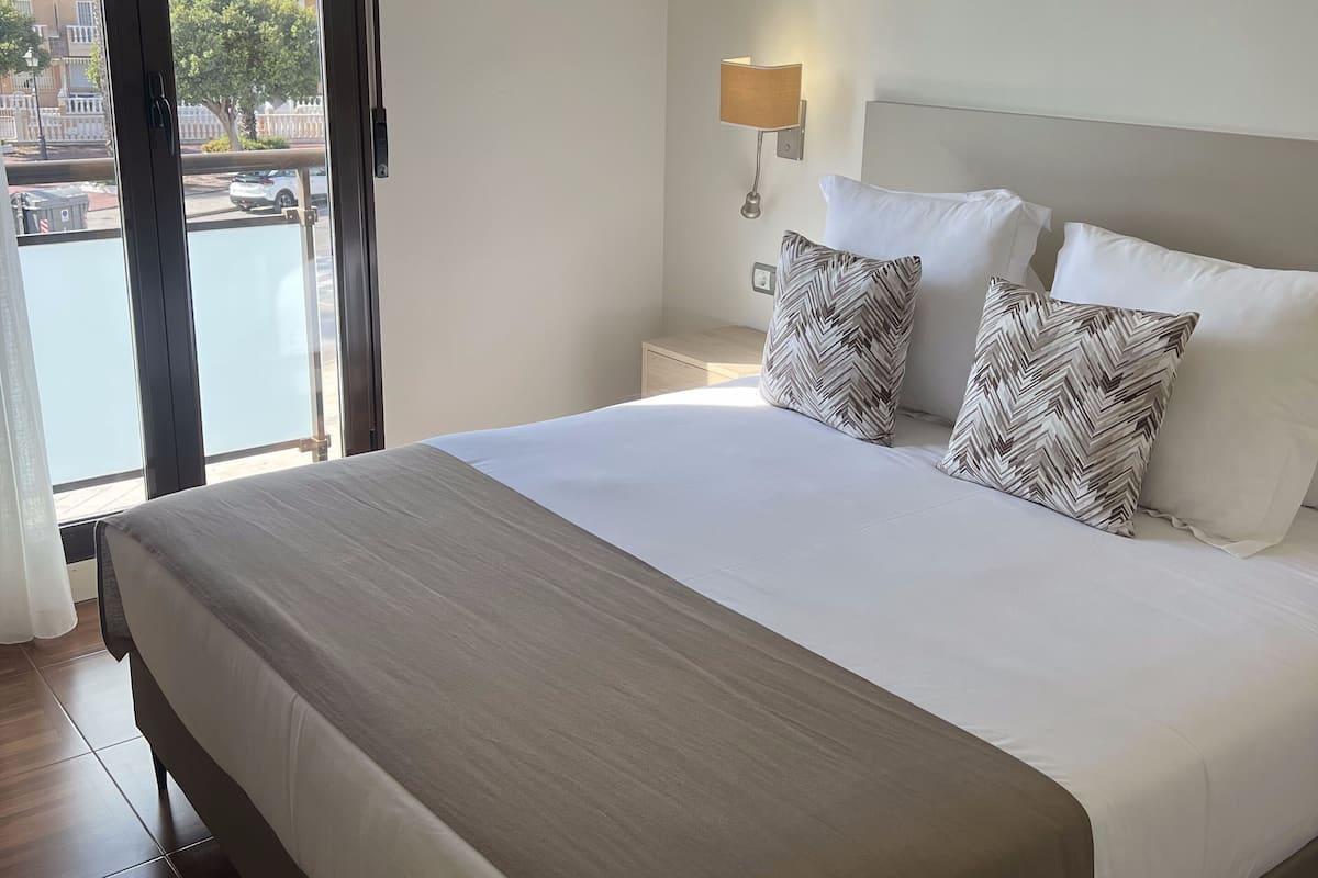 A neatly made bed with a white and beige bedspread, two patterned pillows, a bedside lamp, and a view of a balcony with palm trees.