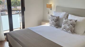Un letto rifatto con ordine, con copriletto bianco e beige, due cuscini decorati, una lampada da comodino e vista su un balcone con palme.