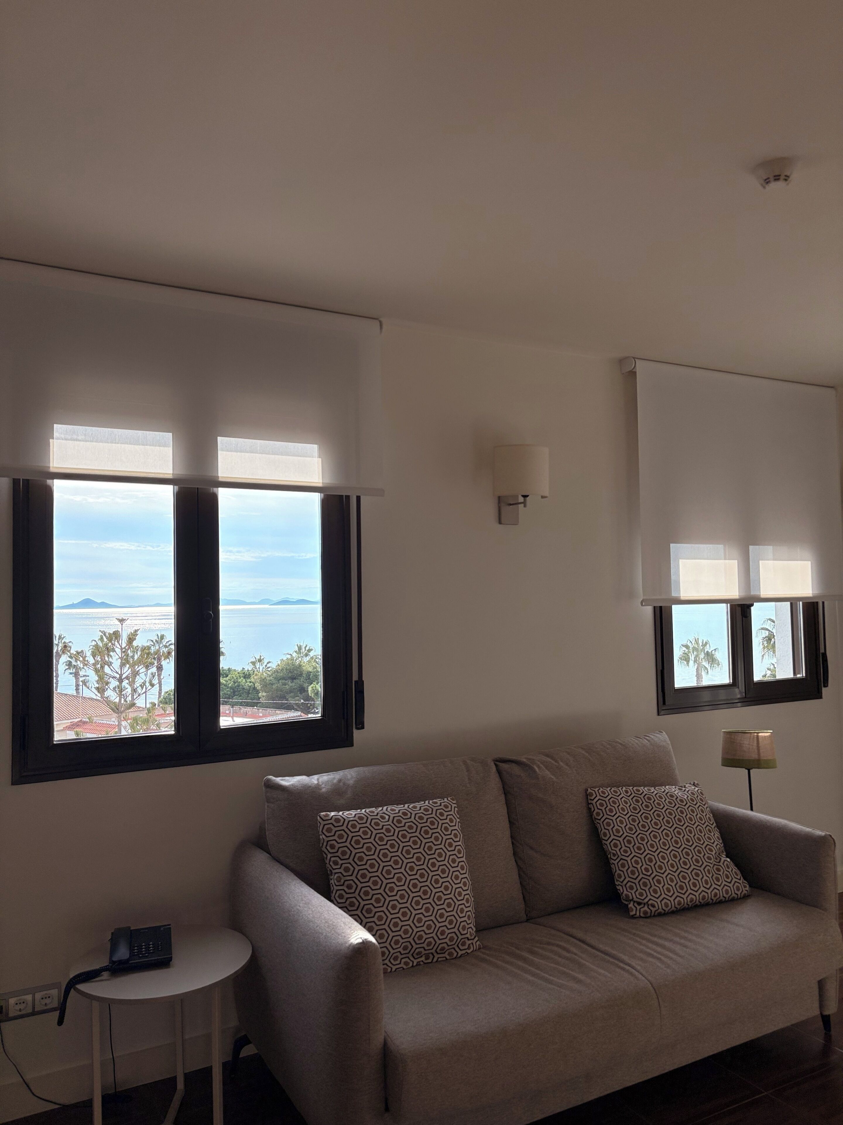 Una sala de estar con un sofá, dos almohadones estampados, una mesita auxiliar con un teléfono y dos ventanas con persianas.