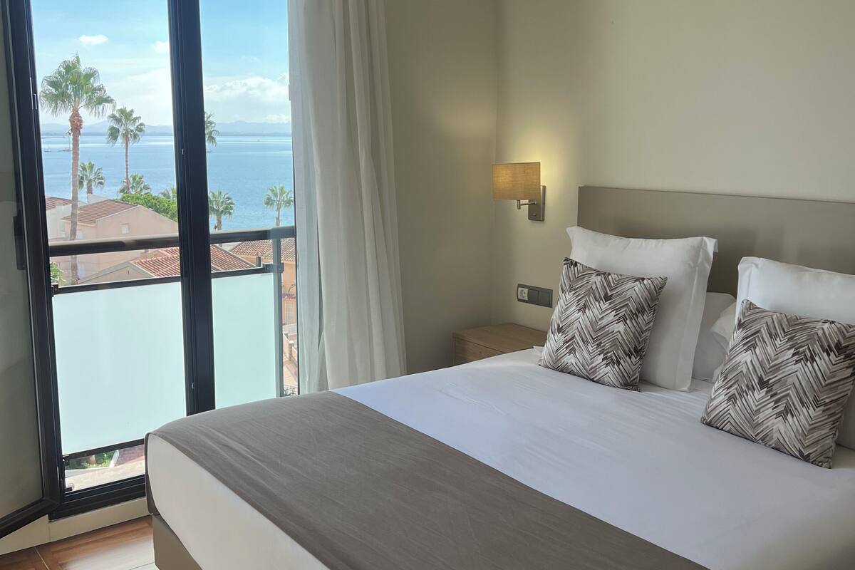 A hotel room with a large bed, a nightstand, a wall-mounted lamp, and a view of the sea through a sliding glass door.