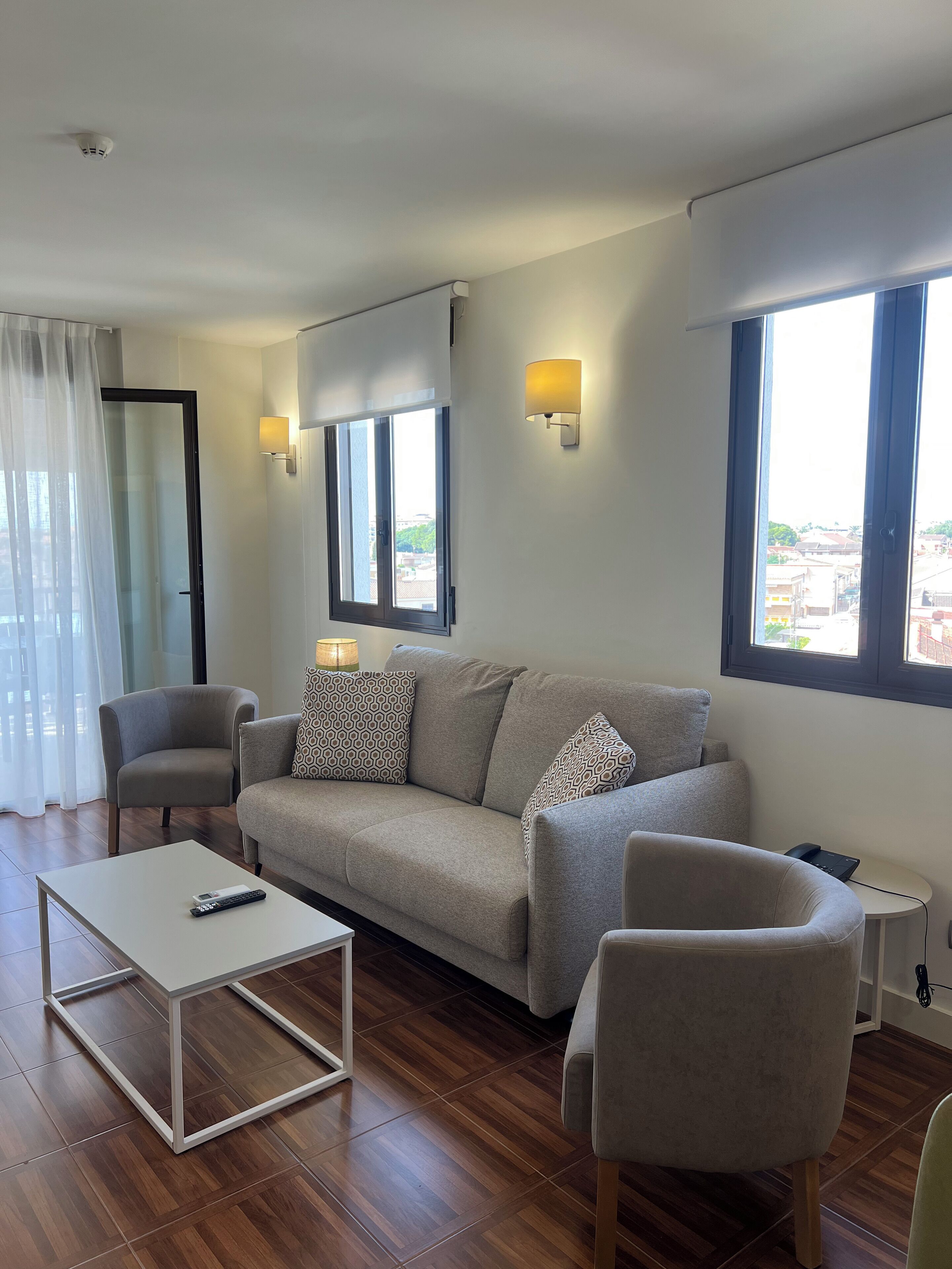 A modern living room with a sofa, armchairs, a coffee table, and a TV. There are two windows with curtains, a wall-mounted lamp, and a ceiling light.
