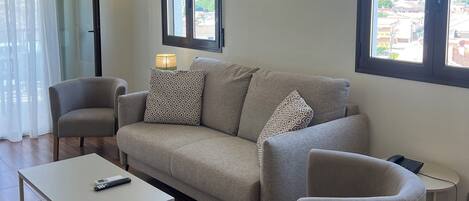 A modern living room with a sofa, armchairs, a coffee table, and a TV. There are two windows with curtains, a wall-mounted lamp, and a ceiling light.