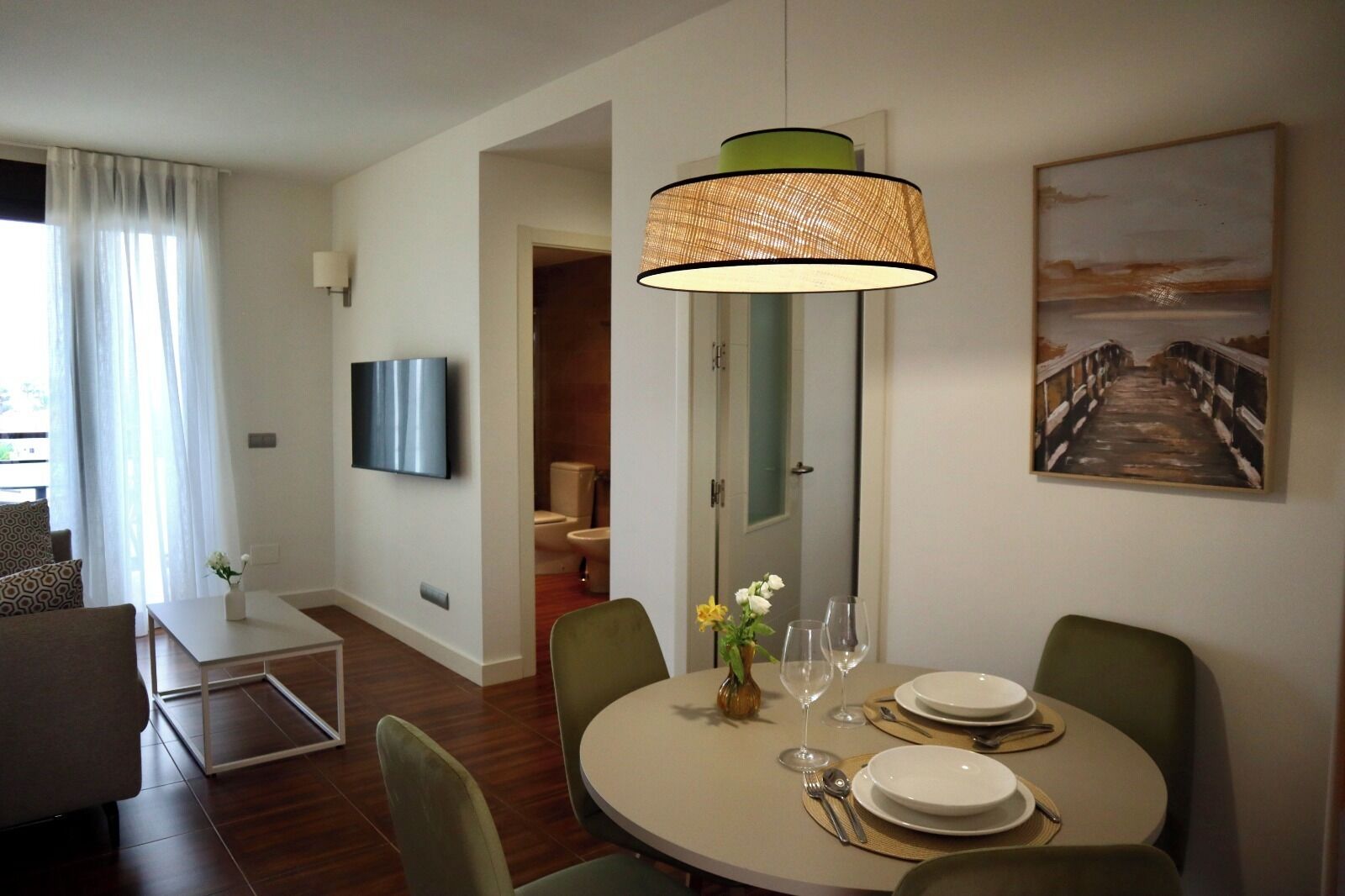 Un espace repas moderne avec une table ronde dressée pour deux personnes, un téléviseur fixé au mur et un tableau au-dessus de la table.