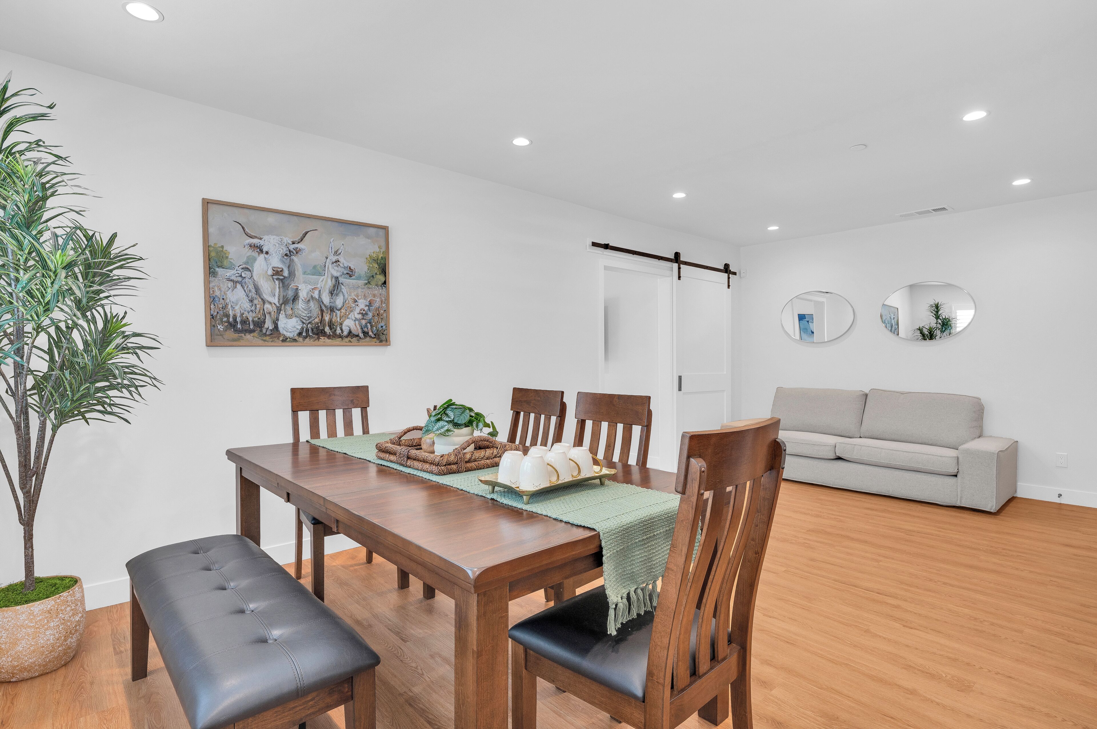 A dining area with a wooden table, chairs, and a bench. There is a painting on the wall, a potted plant, and a sofa in the background.