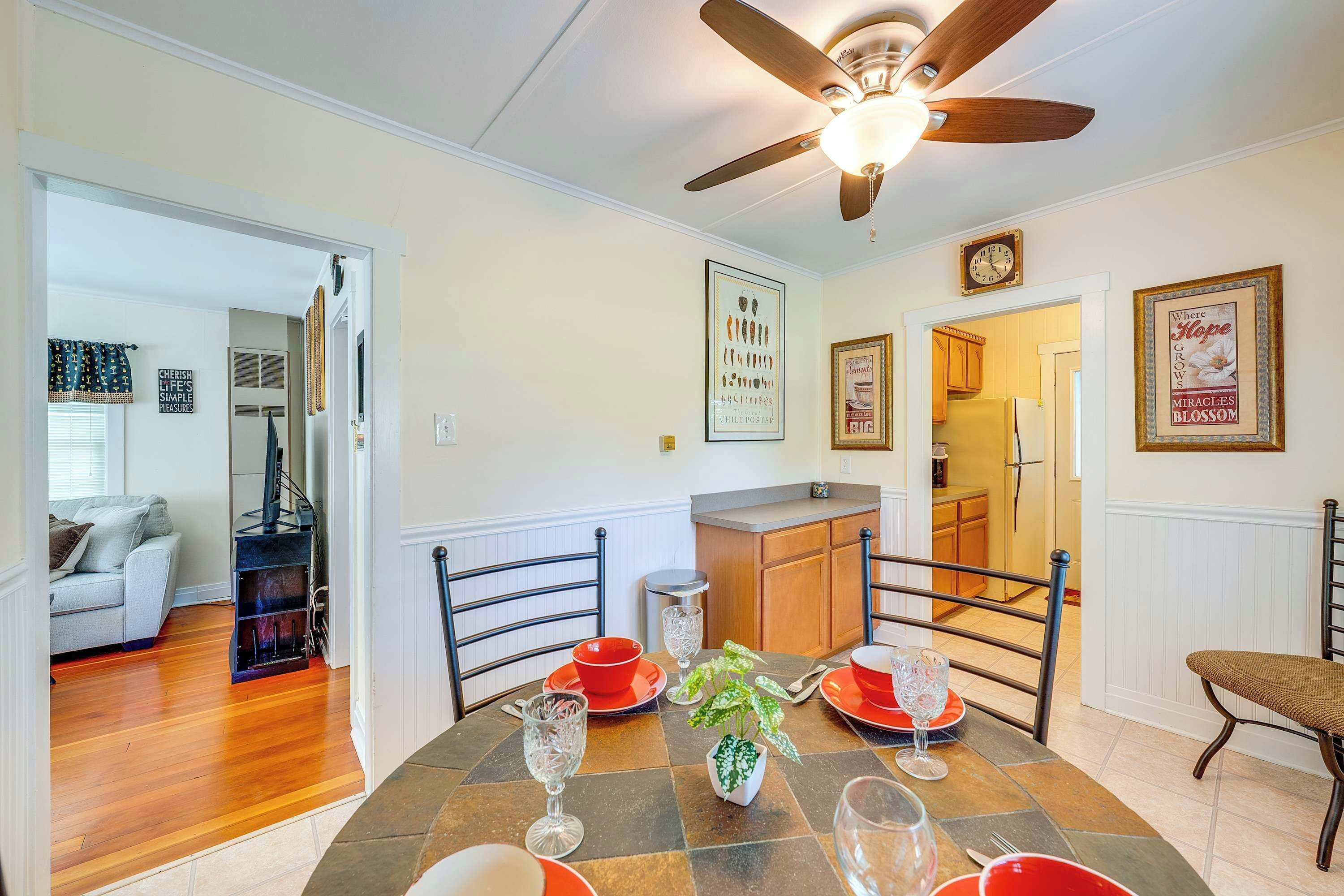A dining area with a table set for four, a ceiling fan, and a kitchen in the background.