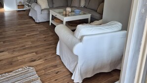 Una sala de estar con sofá, sillón y mesa de centro. Se observa una puerta, estantería en la pared y lámpara colgante.