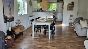 Amplia sala de estar con una mesa de comedor, sillas y un sofá. Se observa una cocina al fondo con una lámpara colgante.