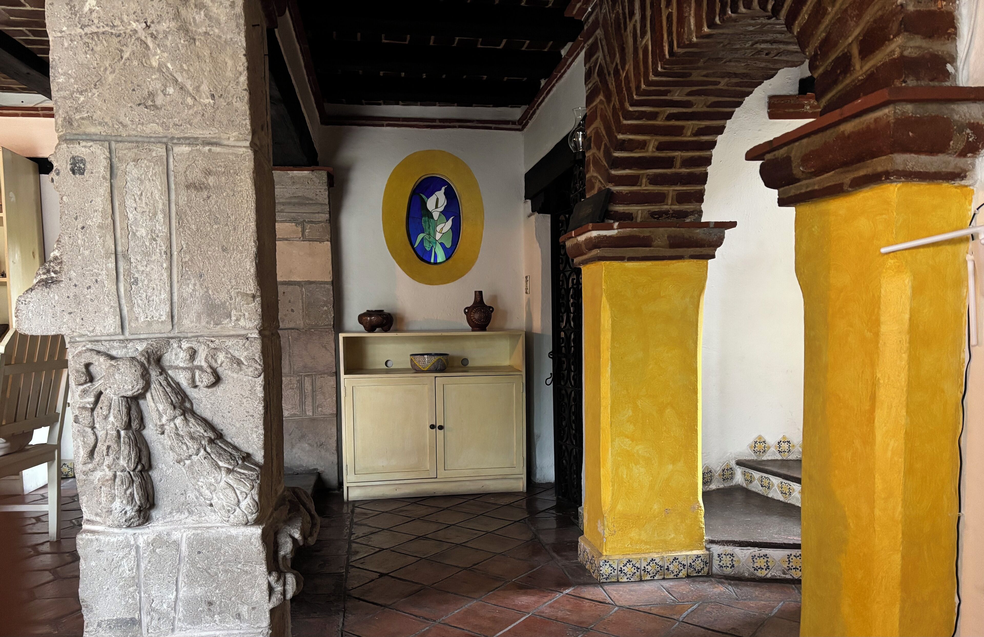 A stone pillar with a carved figure, a yellow wall with a decorative tile border, and a beige cabinet with pottery.