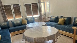 Ein Wohnzimmer mit einem blauen Sofagarnitur, zwei runden Tischen mit weißen Spitzen-Tischdecken und einem Holznachttisch.