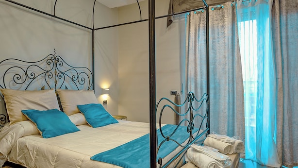 A four-poster bed with a metal frame, a beige bedspread, and blue pillows. There is a small bedside table with a lamp, and a window with sheer curtains. - Luxury Paestum in Naples station train (Napoli)