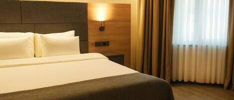 A hotel room with a large bed, a nightstand, a wall-mounted lamp, and a window with curtains.