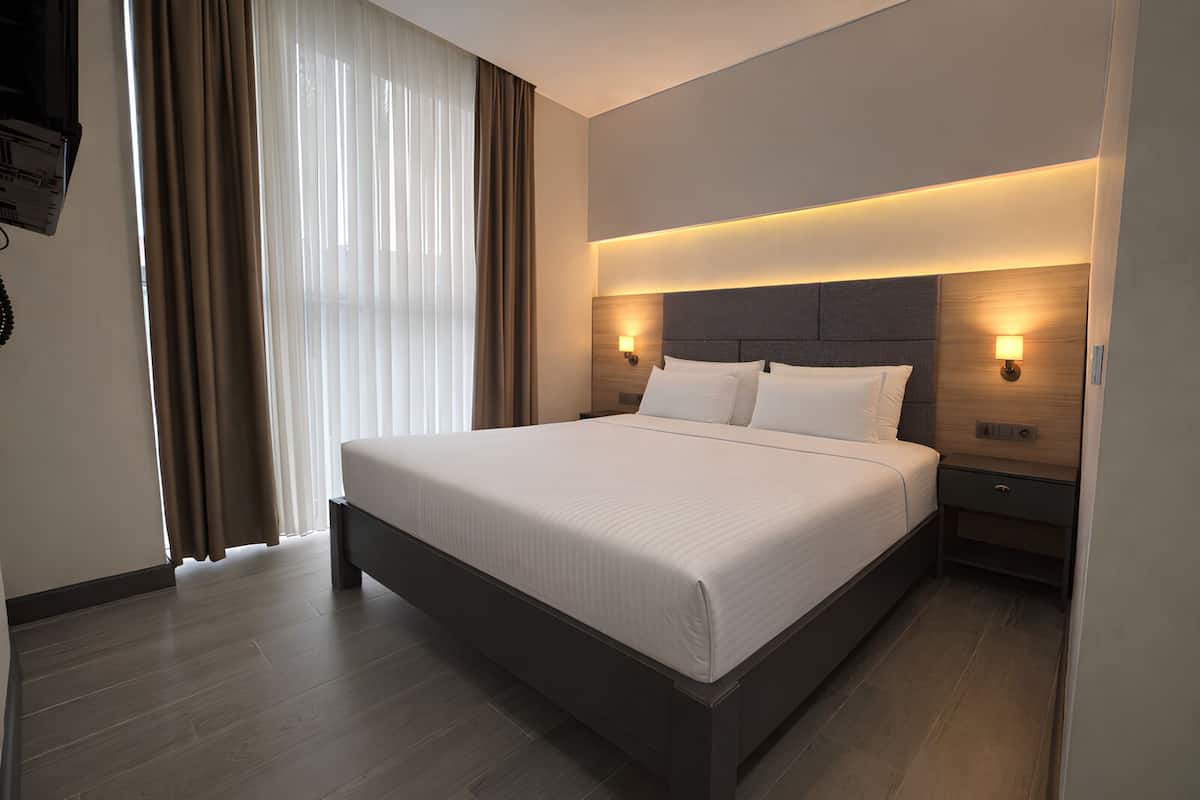 Ein modernes Hotelzimmer mit einem großen Bett, einem hölzernen Kopfteil, Nachttischlampen und Blick ins Freie durch transparente Vorhänge.