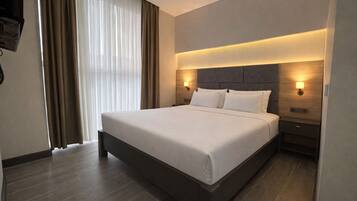 A modern hotel room with a large bed, wooden headboard, two bedside tables with lamps, and a large window with curtains.
