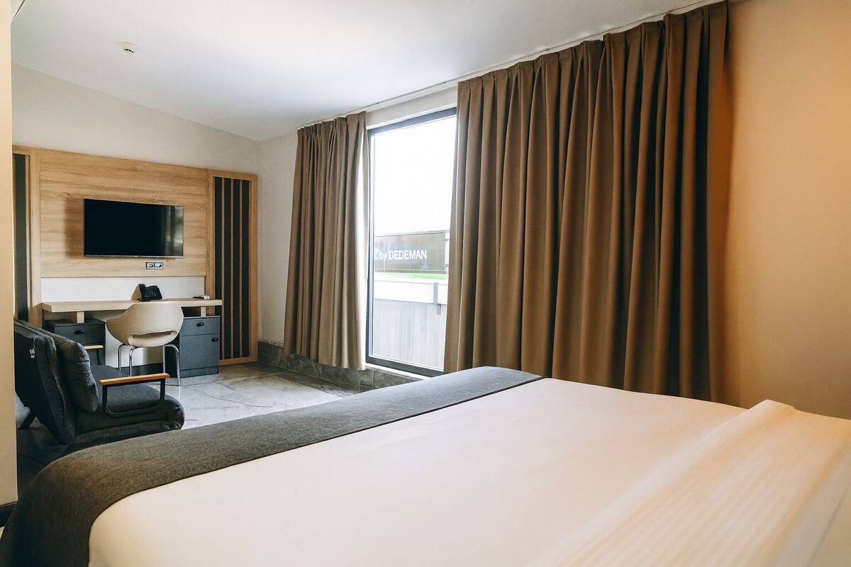Ein Hotelzimmer mit einem großen Bett, einem Schreibtisch mit Stuhl, einem an der Wand montierten Fernseher und Blick ins Freie durch ein großes Fenster.