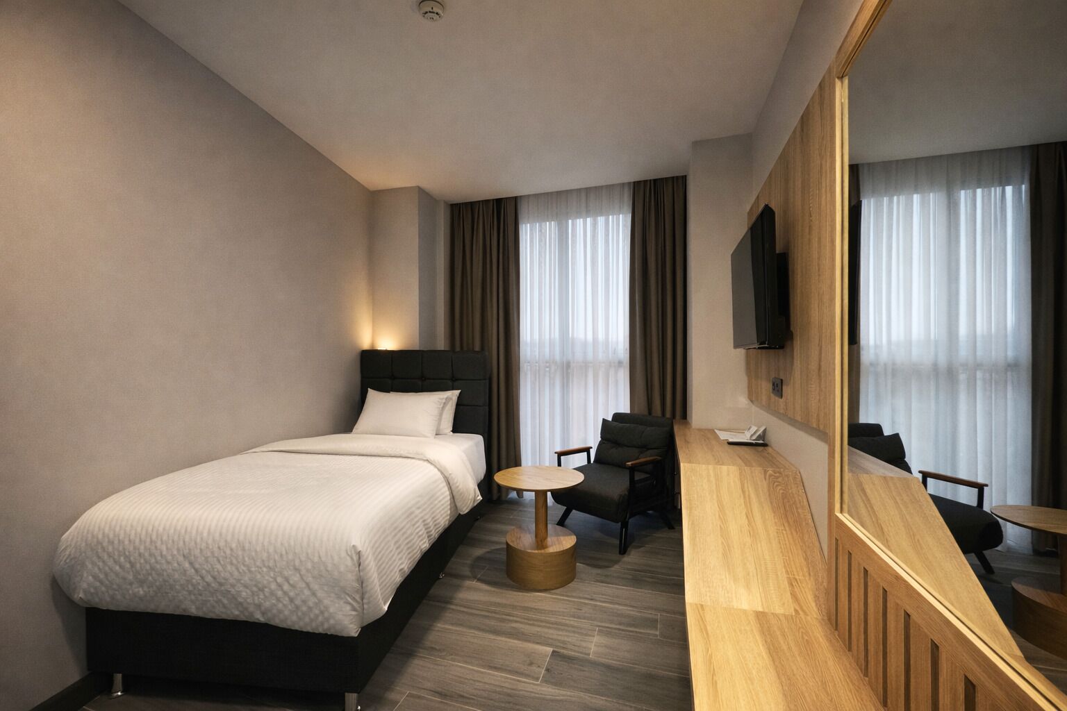 A modern hotel room with a bed, a small round table, a chair, a large mirror, and a TV.