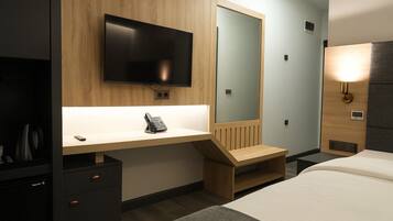 A hotel room with a wooden desk, a flat-screen TV, a bedside table, a bed, and a wall-mounted light.