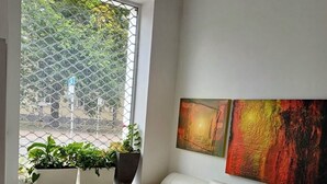 Ein modernes Wohnzimmer mit einem weißen Ledersofa, einem Glastisch und einem großen Fenster mit Blick auf Grünflächen. An der Wand hängen zwei abstrakte Gemälde.