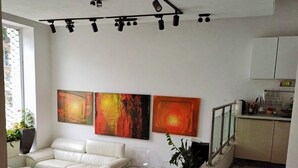 Ein modernes Wohnzimmer mit einem weißen Sofa, einem Glastisch und abstrakten Gemälden an der Wand. Eine Treppe führt zu einer Küchenzeile mit Schränken und einer Arbeitsplatte.