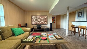 A living room with a wooden floor, a sofa, a coffee table with board games, books, and a gaming console. There is a TV and speakers in the background. - A peaceful, forest retreat with stunning views near historic Harpers Ferry. (Harpers Ferry)