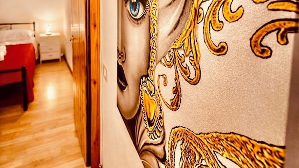 Ein Zimmer mit Holzboden, einem Bett mit roter Bettdecke, einem Nachttisch und einer Wand mit einem großen, verzierten Gemälde.
