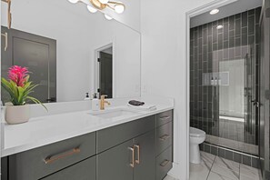 Un baño moderno con un lavamanos blanco, muebles oscuros, un espejo grande y zona de ducha.