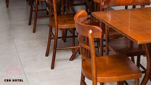 A hotel dining area with wooden tables and chairs, a white handbag on one table, and a logo for CBN Hotel in the bottom left corner. - CBN HOTEL (Parauapebas)