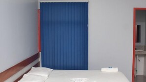 A hotel room with a bed, white linens, a blue curtain, an air conditioning unit, and a bathroom visible through an open door. - CBN HOTEL (Parauapebas)