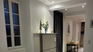 Un coin repas avec une table ronde recouverte d’une nappe à motifs de feuilles, deux chaises, un meuble avec une plante en pot et un escalier menant à une autre pièce.