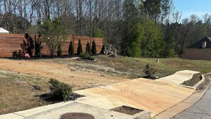 Una strada privata residenziale con cordolo in cemento, un pozzetto e un'area verde alberata con una recinzione in legno.