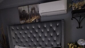 Una camera da letto con una testiera trapuntata, cuscini a righe, una coperta gialla soffice, un comodino con una lampada e un condizionatore d'aria a muro.