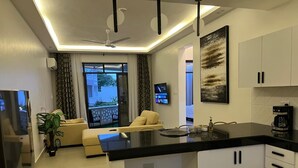 A modern kitchen with a black countertop, white cabinets, and a built-in oven. There is a dining area with a yellow sofa and a TV on the wall. - Nyali Beachfront Apartments Baobab waves (Mombasa)