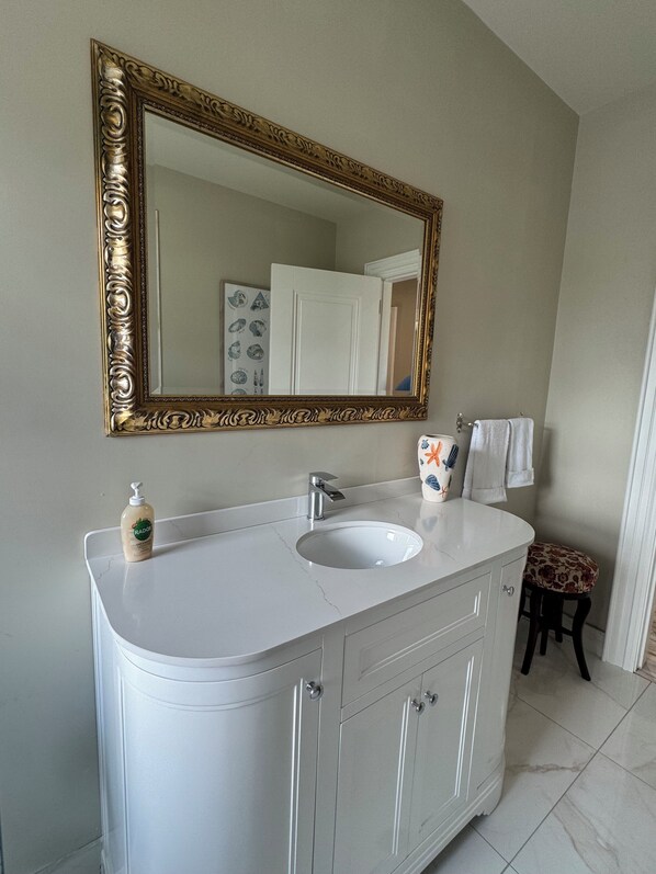 A bathroom with a white vanity, a gold-framed mirror, a white sink, and a small stool with a floral cushion.