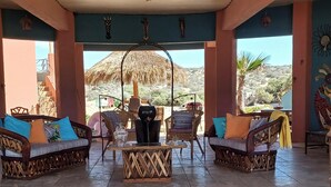 A breezy, open-air lounge with wicker furniture, a central coffee table, and a view of the sea and desert.