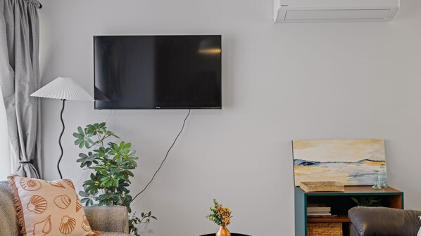 A living room with a flat-screen TV mounted on the wall, a floor lamp, a potted plant, a painting on a shelf, and a sofa with patterned cushions. - The Inlet Hideaway - Inverloch (Inverloch)